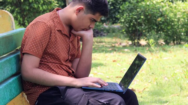 Teenager boy of Indian ethnicity sitting outdoor in nature and using laptop.