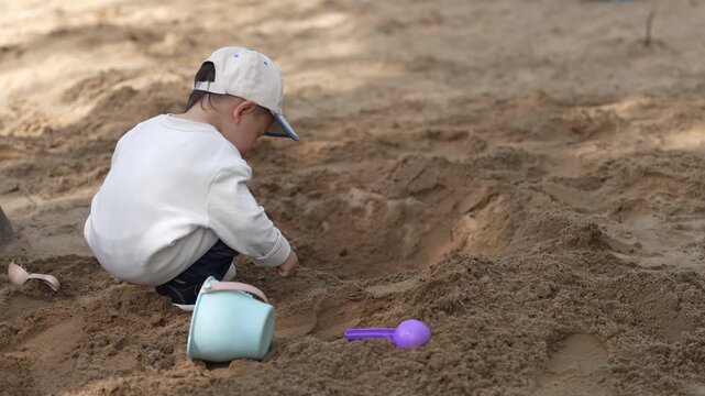 Little Asian Boy Squatting in a Sandbox and Digging Sand Outdoors.