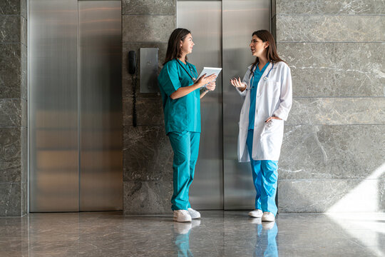 A woman in teal scrubs holds a tablet and discusses with a colleague in a white lab coat at the exit of an elevator in a hospital setting. 