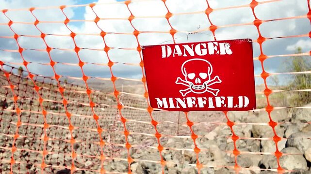 Red sign with a skull and crossbones warning of a dangerous minefield, hanging on an orange safety fence in a desolate, rocky area, indicating a hazardous and restricted military zone