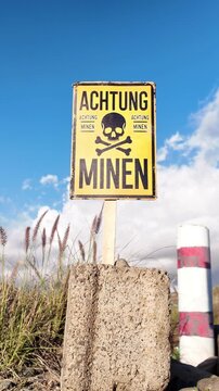 Old yellow minefield warning sign with a skull and crossbones written in German language standing on a concrete block against a blue sky, indicating the presence of deadly explosives