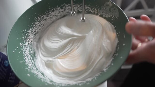 Close-up of egg whites being whipped into a glossy foam with an electric hand mixer in a green mixing bowl.