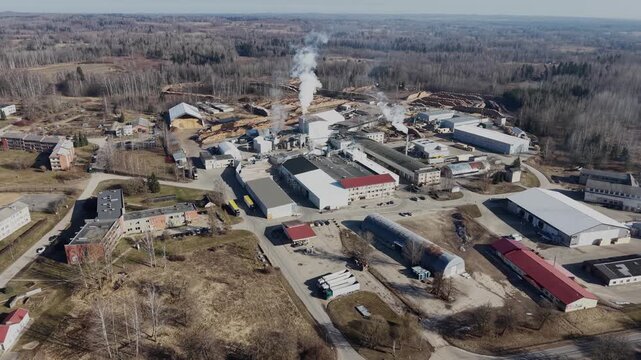 Aerial view of industrial lumber mill with smoke stacks, storage buildings, and logs in a forested landscape during daylight