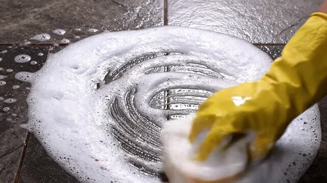 Close up of a person wearing a yellow rubber glove scrubbing a dark textured tiled floor with a sponge and thick white foamy soap bubbles to clean the surface and grout lines in a home.