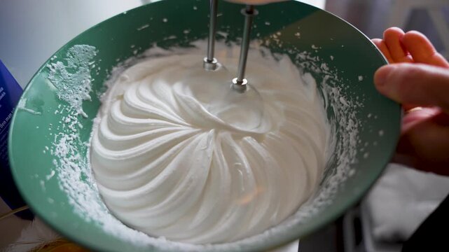 Overhead close-up of an electric mixer whipping egg whites into a glossy spiral pattern inside a green bowl.