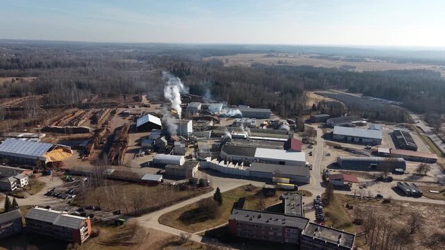 Aerial view of industrial complex with smoke rising from chimneys, surrounded by forest and residential buildings in a rural setting