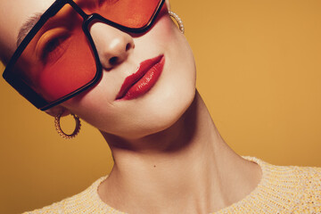 Close-up of stylish woman wearing oversized red sunglasses and gold hoop earrings on yellow background. Fashion portrait with bold makeup and trendy accessories. © SHOTPRIME STUDIO