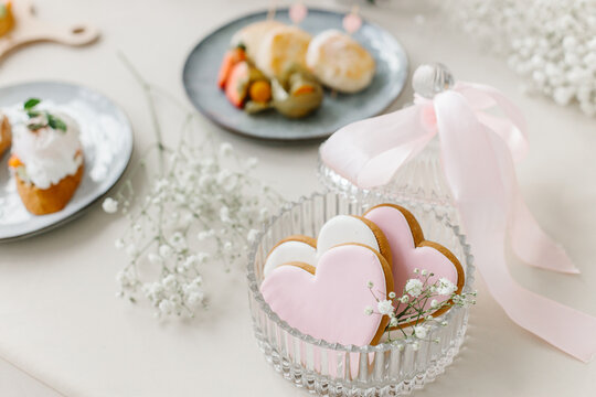 Sweet pink heart cookies in a crystal jar with a pink ribbon set on a festive table with blurred appetizers behind.