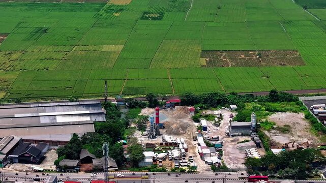 Aerial view of an industrial concrete batching plant and cement mixer trucks parked next to lush green rice fields in rural Indonesia.