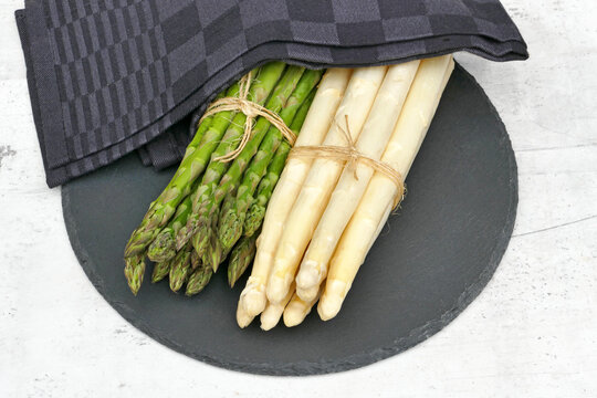 Gr&uuml;ner und wei&szlig;er Spargel, jeweils zu B&uuml;ndeln gebunden, liegen auf einer runden schwarzen Schieferplatte unter einem dunkelgrauen K&uuml;chentuch.