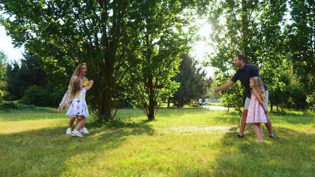 Positive family throwing frisbee disk in park. Smiling girl catching frisbee plate. Young parents playing frisbee with daughters. Family enjoying summer outdoor activity. Happiness