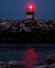 A small red lighthouse beacon glows over a rocky breakwater at dusk, reflecting vivid red light across the calm dark water beneath a deep blue evening sky. © Aperezer