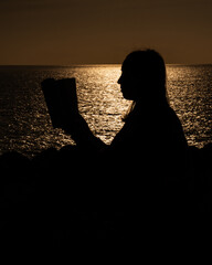 Silhouette of a woman reading a book by the sea at sunset, with warm golden light shimmering across calm water, creating a peaceful and contemplative mood. © Aperezer