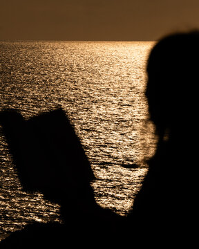 A solitary silhouette holding a book faces a shimmering sea at sunset, warm golden light reflecting on gentle waves, creating a calm, introspective coastal mood.
