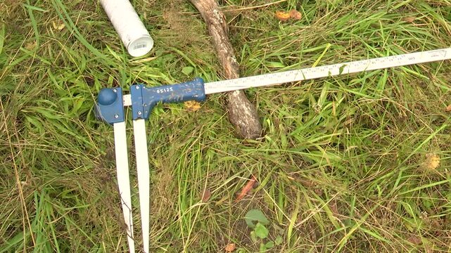 Measuring tool positioned on grass with a stick and a white tube nearby, showcasing the gradual adjustment of the measuring arms in a natural outdoor setting