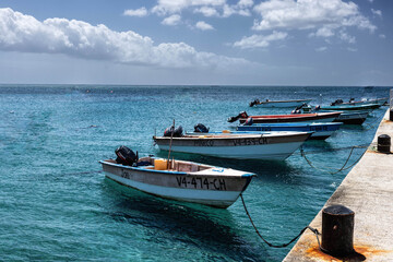 Naklejka premium This serene harbor scene in St. Kitts features several small fishing boats floating calmly in the vibrant turquoise Caribbean Sea, moored to a concrete pier on the right. 
