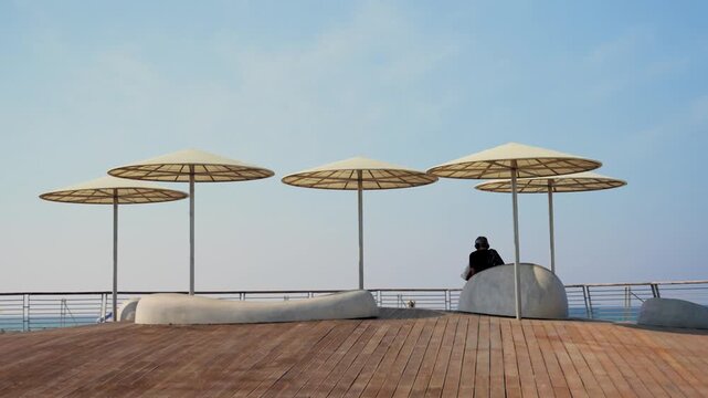 Man sitting with headphones in Tel Aviv beach promenade under sun umbrella. Sunny day in Israel. Wooden deck and sun shades overlooking Mediterranean Sea. Tourist or local enjoys sitting alone