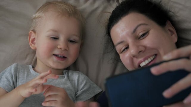 Happy mother and little son lying in bed making video call to relatives. Cute toddler is smiling, talking and waving hands at smartphone screen, enjoying connection with father. Family life with kid