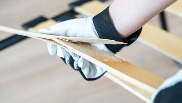 Hand holds wood plank with care and precision. White glove protects fingers from splinters or dust. Background shows wooden frame assembly in progress