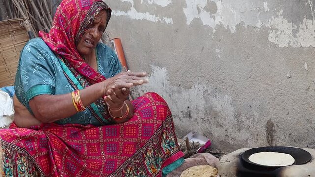 An elderly Indian village woman in traditional attire cooking handmade rotis (flatbread) on a traditional mud stove (chulha) using firewood in a rural kitchen.
