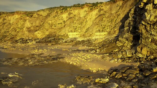golden cliff coastline at sunset with shallow rockpools and reflective wet sand, layered strata and scattered pebbles, tranquil mood for nature editorials