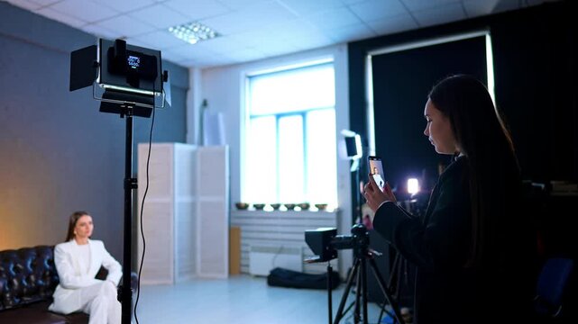 Photo studio interior with a filming process. Man and woman work backstage recording a woman in white suit on the sofa.