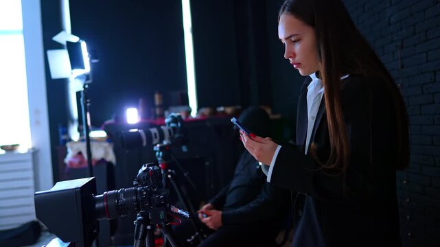 Cameras on tripods and soffits are set in photo studio. Male photographer and female assistant are near the equipment backstage.
