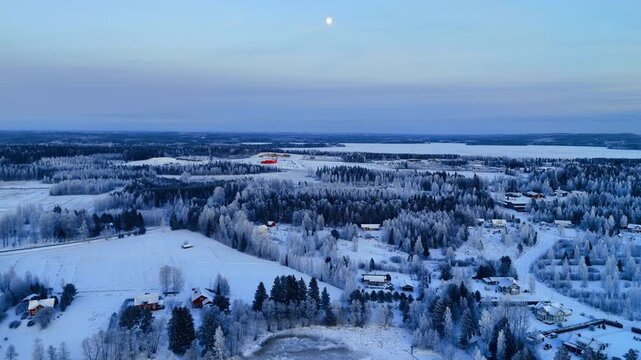 Aerial view of snow covered village in Lapland winter. Aerial shot of houses, frosted trees, and frozen lake under a pale blue sky at twilight in Lapland.