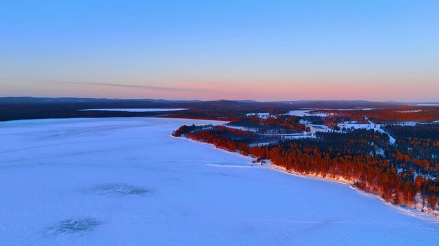 Aerial view of snowy forest edge along a frozen lake. High angle shot of trees casting long shadows on snow during a winter sunset in Lapland.