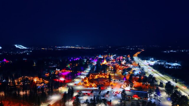 Aerial perspective of snowy road and illuminated village at night. Aerial view showing a long road leading to a brightly lit arctic village during winter night.