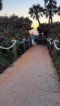 Sandy path to the beach at sunset in Florida. A sandy walkway with rope fencing leading to the ocean during a vibrant sunset with palm trees.