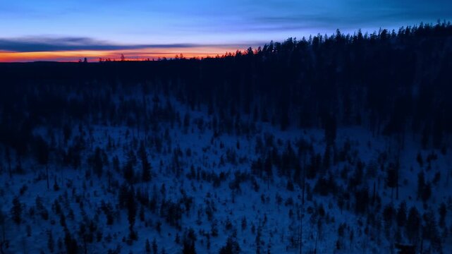 Twilight aerial view of snowy hilly forest landscape in Lapland. Low light aerial shot of a dense snowy forest on hills in Lapland during blue hour twilight.