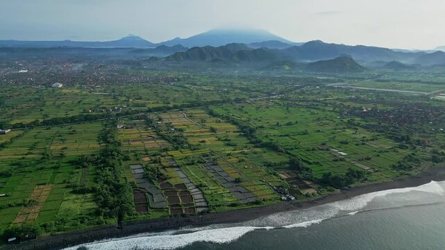 Aerial farm grid volcano Agung patchwork rice paddies and irrigation pools, waves lapping pebble shore, distant misty peaks, agricultural mosaic stretching toward coastline,Bali,Indonesia