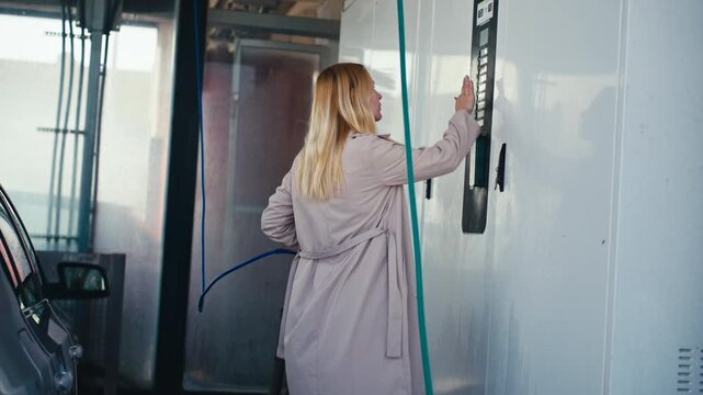 Woman in trench coat inserting payment card into machine at self-service car wash, activating system, picking up high-pressure water sprayer.Fashion meets routine work.