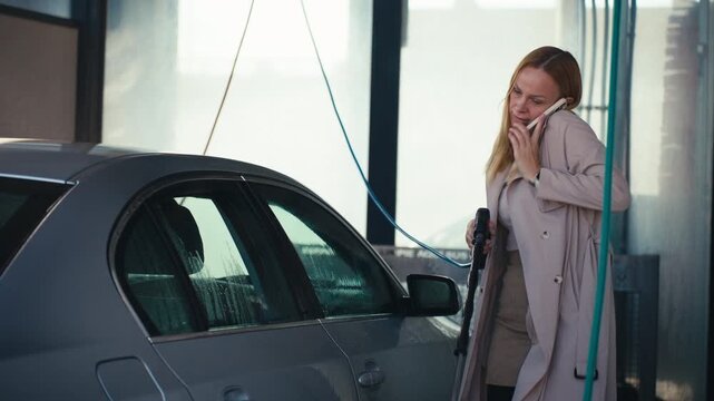 Multitasking businesswoman simultaneously wash sedan with high-pressure hose and talking on smartphone at self-service car wash station during hectic day.Concept independence