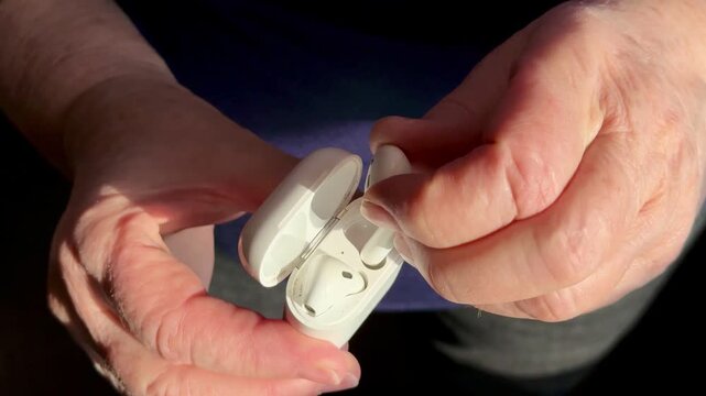 Closeup woman holding white earbud case, gently removing single earbud for listening warm sunlight highlights textured hands.