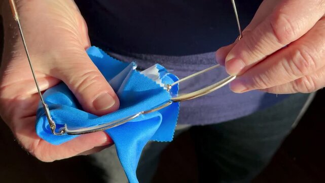 Closeup hands cleaning eyeglasses with satin cloth, woman gently polishes gold frames and lenses, sunlight highlights microfiber.