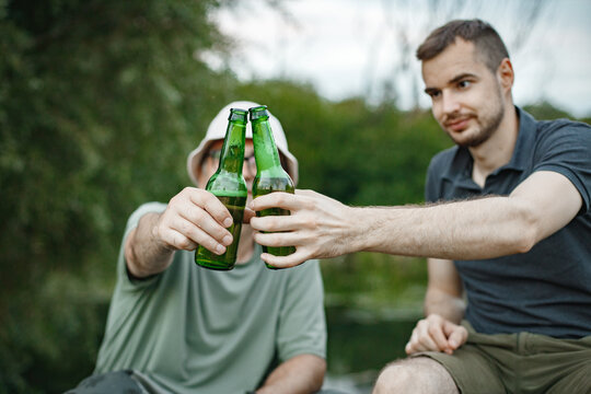 Cropped photo of a men near the lake clinking with two bottles of beer