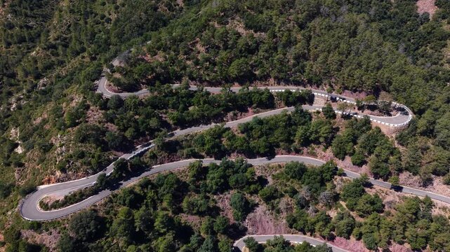 Aerial view of cyclists descending a serpentine mountain road with sharp hairpin bends through dense green forest in Sierra de Espadan, Castellon, Spain