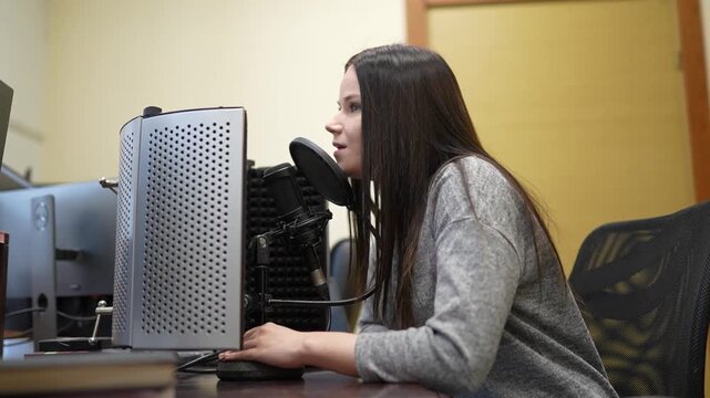 Young woman with long hair records audio in a studio, using a microphone and computer equipment in a professional setting