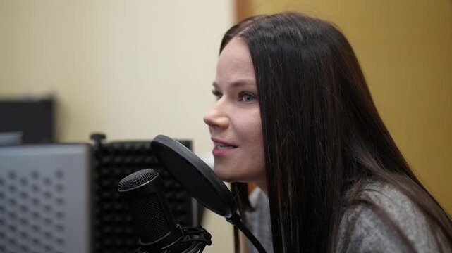Female voice artist with long dark hair speaks into a microphone in a recording studio, soundproof panels and equipment visible in the background