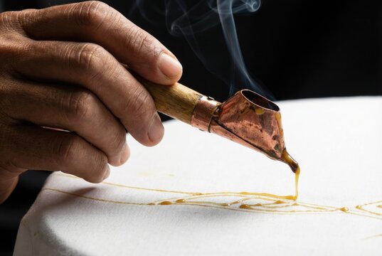Hand using a traditional copper tool to draw lines of hot molten wax onto white fabric