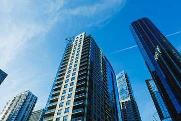 Fototapeta premium Tall buildings reach for the sky in a modern city under clear blue skies with wispy clouds and bright sunlight reflecting off the glass surfaces