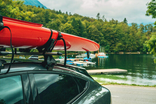 Closeup of red kayak mounted on car roof rack, parked by Lake Bohinj in Slovenia, with pier, boats, and green alpine forest in view.
