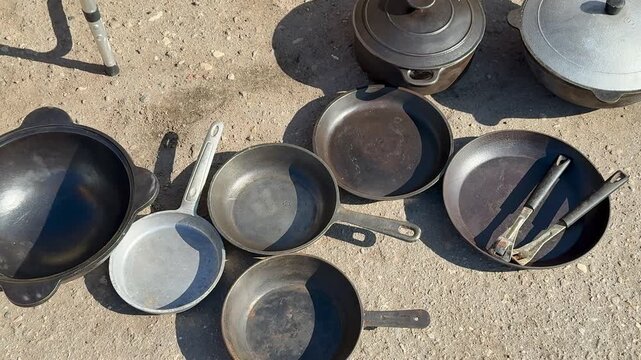 Worn pans pots cauldron on asphalt, sunlit display of used cookware for sale, cast iron skillets and scratched lids scattered.