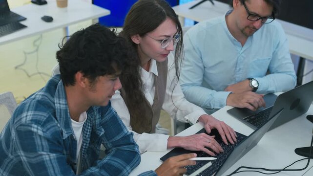 collaborative AI development team in modern tech office: young male developer coding on laptop, female team leader in light blue blazer pointing at code and neural network visualization	