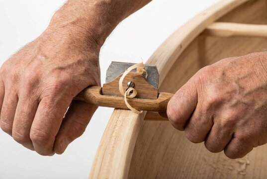 Boatbuilder hands using sharp wooden spokeshave tool to shape and smooth cedar wood on canoe hull