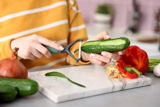 Woman peeling fresh cucumber at grey table with vegetables indoors, closeup