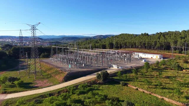 Overhead view of a high voltage electrical substation with transmission towers and power lines set among green pine forest hills and mountains under clear blue sky