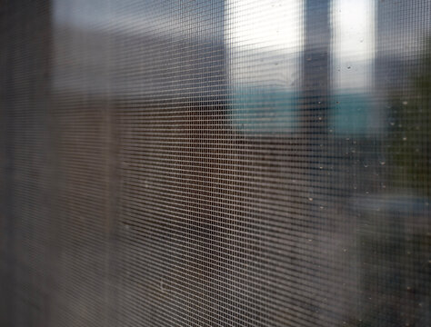 Close up perspective of a grey fiberglass window insect screen mesh showing an intricate woven grid pattern with a heavily blurred outdoor scenery visible directly behind
